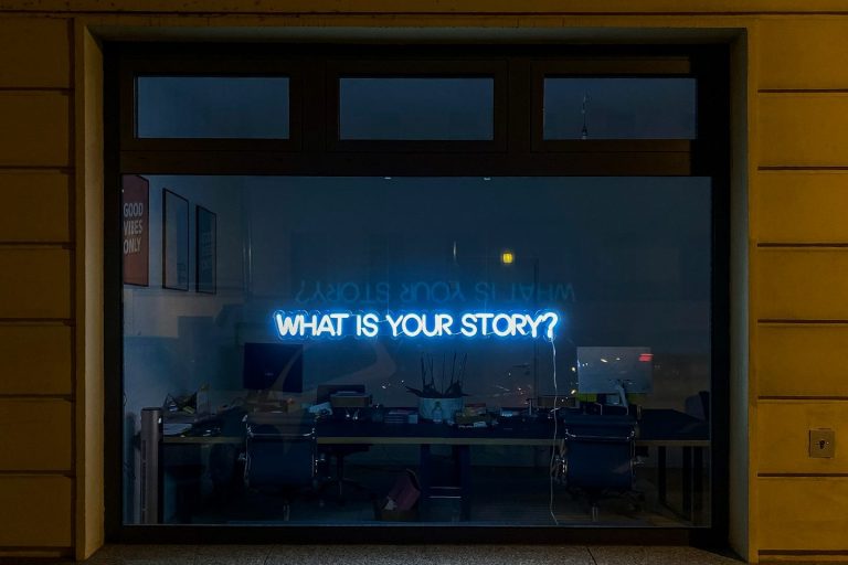 Neon sign in a window displaying the text "What is your story?"
