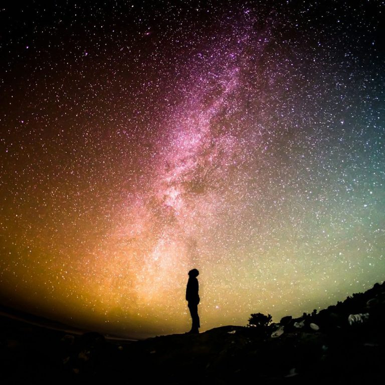 A silhouetted figure stands under a vibrant Milky Way galaxy in a night sky.