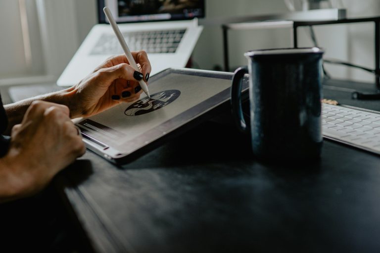 A person drawing on a tablet beside a mug and a laptop on a desk.